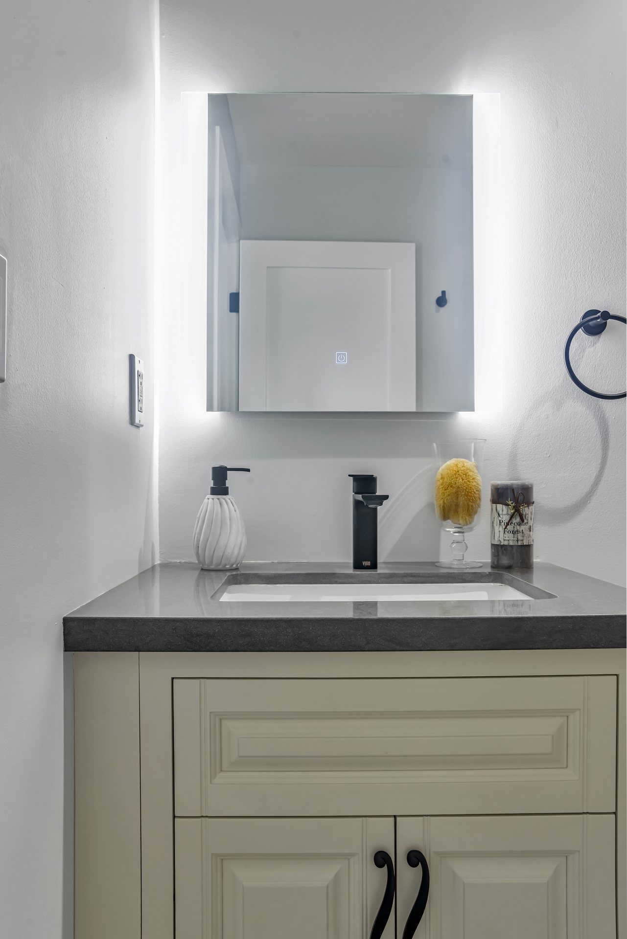 Cream raised-panel vanity with a gray countertop, black faucet and backlit mirror